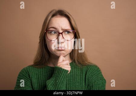 Thoughtful young woman with hand on chin looking up, expressing doubt, confusion, or hesitation on brown background. Stock Photo