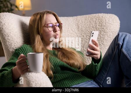 Young woman with glasses reacts emotionally while looking at smartphone screen, holding a coffee cup at home. Stock Photo
