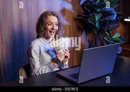 Excited woman holding cash, celebrating online success, winning money, financial achievement, remote work earnings, joyful reaction at laptop Stock Photo