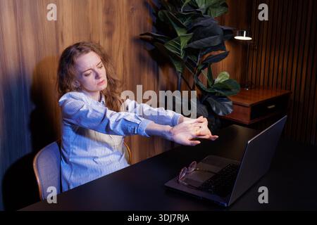 Woman stretching hands while working on a laptop, relieving tension, taking a break from remote work in a home office. Stock Photo