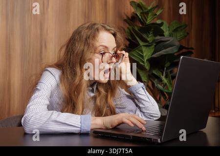 Shocked young woman with glasses looking at a laptop screen, expressing surprise, disbelief, or amazement while working or browsing online. Stock Photo