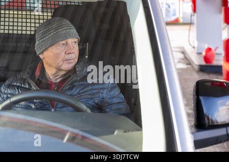 Elderly man in a beanie is seated in a car at a gas station, gazing out the window, creating a serene atmosphere of thoughtfulness and daily routine Stock Photo