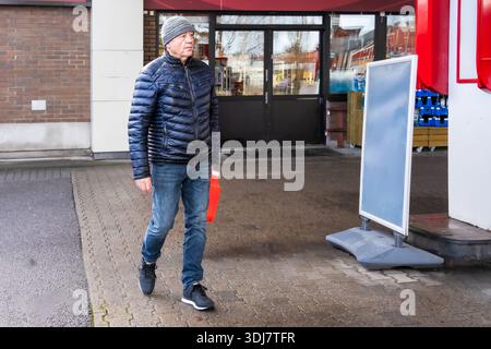 Elderly man in blue puffer jacket and gray beanie walks through a commercial space, holding a red bag, capturing the essence of urban daily life Stock Photo
