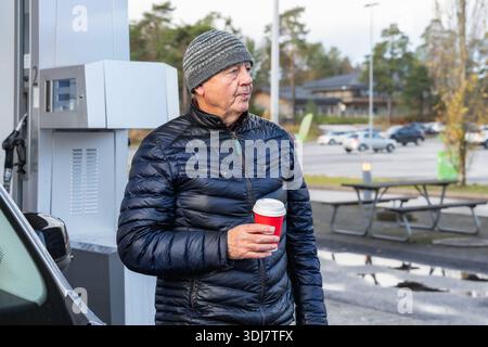 Elderly man in a puffy jacket and beanie stands at a gas station, holding a coffee cup, with vehicles and greenery in the background creating a calm a Stock Photo
