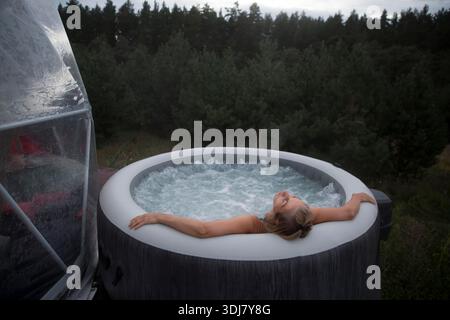 middle aged woman rests in a hot tub in autumn cold weather against the background of green pine forest. Weekend getaway at glamping. || Model released Stock Photo