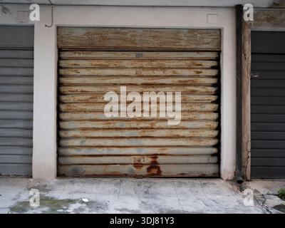 A rusted metal roll-up garage door showing signs of long-term neglect. The door is heavily corroded, with visible rust streaks. The surrounding white- Stock Photo