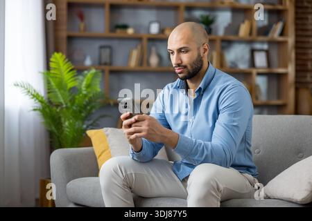 Young man relaxing on couch browsing internet on mobile phone, staying connected with digital technology, social media, and online communication in a comfortable living room Stock Photo