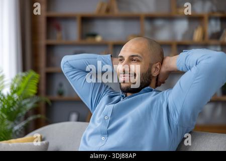 Middle eastern man sitting on a sofa with hands behind his head, smiling and looking away, enjoying a peaceful break from work in his comfortable living room Stock Photo