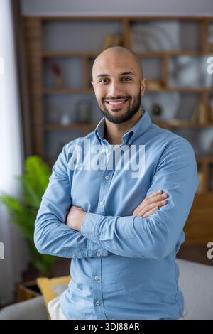 Smiling bald bearded man in a blue shirt stands with crossed arms in a warm home interior, confident and relaxed, looking at camera in a casual professional portrait Stock Photo