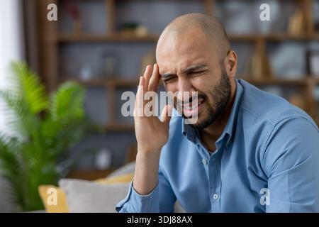Young man experiencing intense headache, migraine, or stress, holding his head while grimacing, expressing discomfort and pain from mental tension indoors Stock Photo