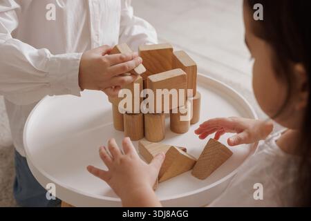 Children playing with wooden blocks, building a tower together on a white table, engaging in creative and educational play in a cozy home setting. Stock Photo