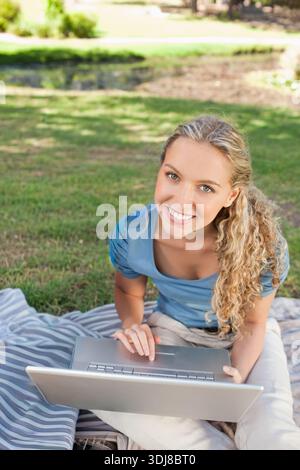 Woman sitting on striped picnic blanket on park lawn using silver laptop, wearing blue top. Outdoors, nature, sunlight, pond, trees, greenery, serenit Stock Photo