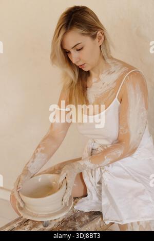 Young female artist shaping a handmade ceramic bowl, covered in clay, in a rustic studio with soft, natural lighting. Stock Photo