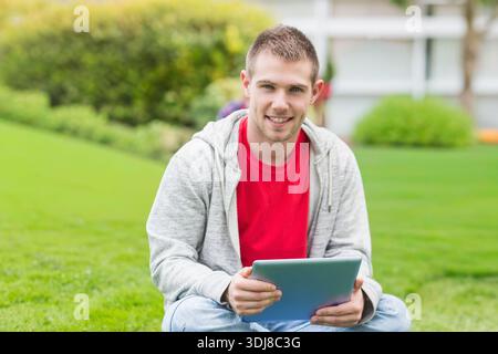 Man sitting on grassy lawn holding tablet, wearing red tee and gray hoodie, smiling at camera. Adult, male, device, campus, outdoor, lifestyle, bright Stock Photo