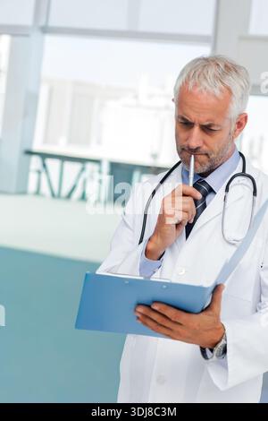 Senior male doctor reading blue clipboard, in lab coat with stethoscope at clinic, copy space. Physician, healthcare, medical, office, windows, bright Stock Photo