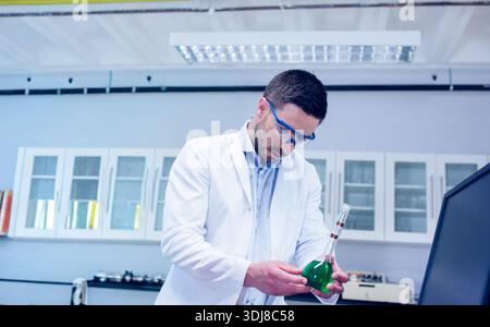 Male scientist in white lab coat and blue goggles holding green flask and inspecting at bench. Laboratory, technician, liquid, monitor, research, chem Stock Photo