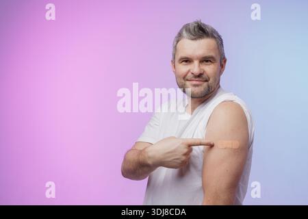 Happy young adult indian hispanic man showing patch on arm after vaccination isolated on pink blue background. modern vaccination and immunization of Stock Photo