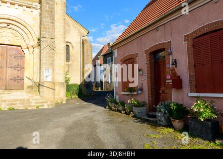 Unknown location - June 28, 2022: The left side of a stone church with an arched doorway, ornate wooden doors, metal handrails, and a large star decoration is visible next to a pink residential house with closed shutters, potted plants, and a red mailbox on a quiet village street under a blue sky. Stock Photo