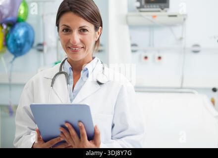 Mid-adult female doctor standing in patient room with lab coat, tablet, stethoscope, balloons. Clinician, hospital, medical, device, headshot, celebra Stock Photo