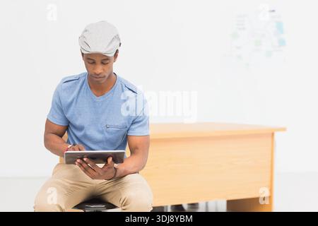 Adult African American man sitting on stool using tablet in bright studio wearing flat cap. Minimal, modern, white, desk, device, map, headwear Stock Photo