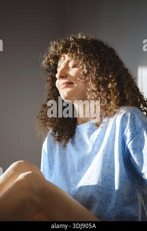 A woman sits relaxed by a window with sunlight on her face. She has curly hair and wears a blue shirt. The light creates soft shadows around her as sh Stock Photo