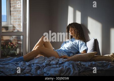 Young woman sits on a bed in a room with sunlight streaming in from a window. She has curly hair and wears a light blue pajamas and socks Stock Photo