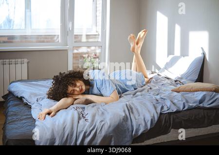A young woman lies on her stomach on a bed, wearing blue pajamas. Sunlight comes through the window and shine her feet. Plants are visible outside. Th Stock Photo