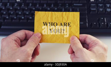 A person holding a yellow sticky note with the question WHO ARE YOU written in on it against a computer keyboard background. Close up. Stock Photo