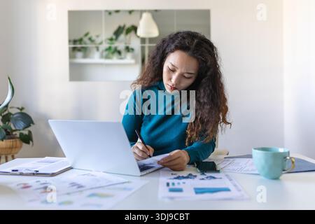Focused Asian woman using laptop at home, looking at screen, chatting, reading or writing email, sitting on couch, female student doing homework, work Stock Photo