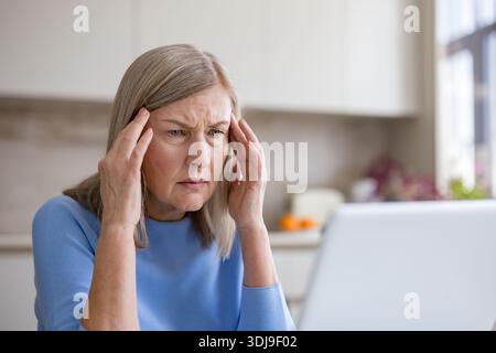 Senior woman with white hair holding her temples in pain, frustrated and stressed while working on a laptop at home, struggling with a headache or migraine and concentration issues Stock Photo