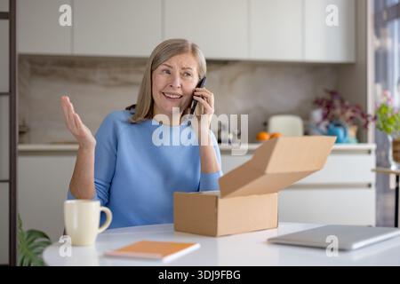 Senior woman is upset, talking on a smartphone, holding her hand up, and looking into an open delivery box, expressing frustration about a potential issue with her online purchase Stock Photo