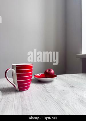 A vibrant red ceramic cup with a white striped pattern stands next to a ripe red apple on a saucer, arranged on a light-colored wooden surface in a cl Stock Photo