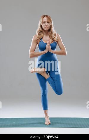 Young woman practicing tree pose on yoga mat, maintaining balance and focus in a studio wearing blue outfit. Stock Photo