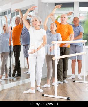 Experienced mature female ballet dancer, dressed in sportswear and group of women and men engaged in classical ballet performs exercise standing in Stock Photo