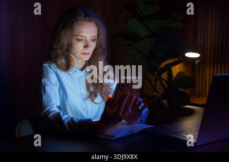 Woman sitting at night on her smartphone, at her desk at home, night work. Stock Photo