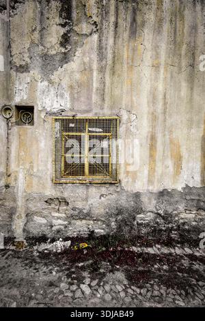 An aged wall with a weathered window featuring a metal grill, showcasing rustic textures and colors in an urban environment Stock Photo