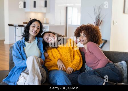 Diverse female friends sitting on gray sofa in modern kitchen, wearing denim and yellow sweater Stock Photo
