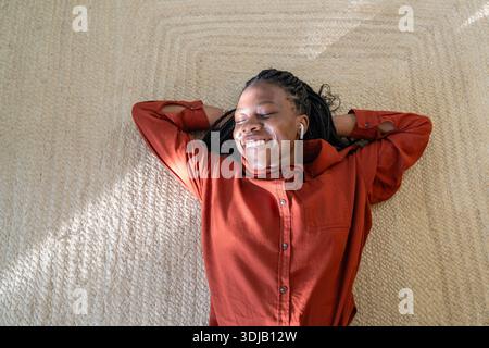 Carefree happy African American woman lying on floor relaxing listening to favorite music in pods. Stock Photo