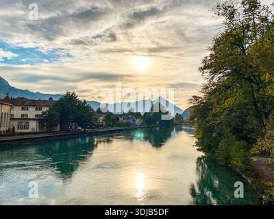 A scenic evening view looking down the Aare River in Interlaken, Switzerland. The image captures the sun setting behind the distant Swiss Alps (resemb Stock Photo