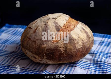 A crusty round loaf of sourdough rests on a blue plaid kitchen towel, evoking artisanal baking, warmth, and homemade comfort. Stock Photo
