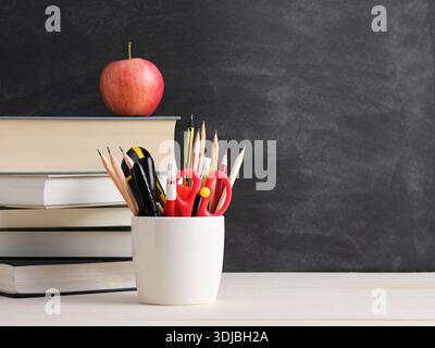 Stack of books with a red apple on top, a cup holding pencils and scissors, set against a blackboard background, symbolizing education and learning Stock Photo