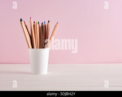 Collection of vibrant colored pencils neatly arranged in a white cup against a pastel pink background. Stock Photo