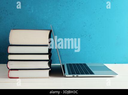 Stack of books and a laptop symbolizing online education and e-learning placed on white wooden desk in front of blue background Stock Photo
