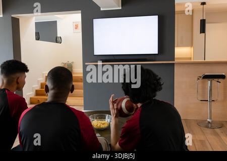 Diverse male friends in red-black shirts watching TV on sofa holding football near bowl of chips Stock Photo