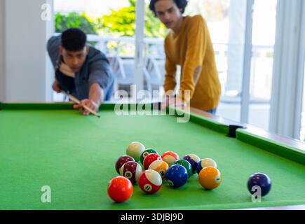 Two men lining up shot on green-felt table near glass doors with cue stick, racked balls Stock Photo