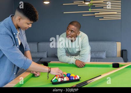 Diverse male friends racking billiard balls into triangle rack on green pool table in lounge Stock Photo