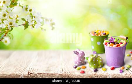 A cheerful spring setup featuring candy-filled pastel buckets, ceramic bunny figurines with polka dots, and blooming white blossoms in the background. Stock Photo