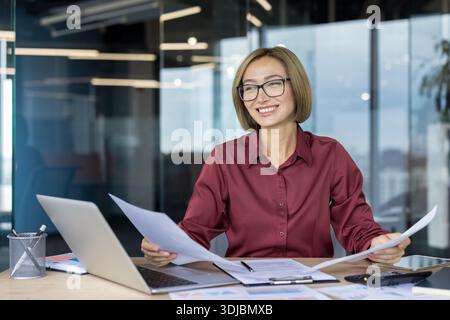 Smiling young businesswoman confidently analyzing paperwork and financial data at her desk, working efficiently in a bright and modern office environment Stock Photo