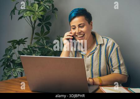 A cheerful non-binary individual with vibrant blue hair sits at a wooden desk, engaging in a phone conversation while working on a laptop in a modern, Stock Photo