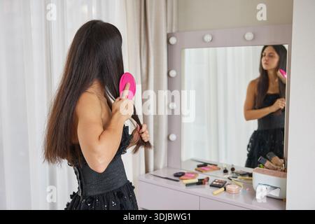Woman standing in front of illuminated mirror, brushing her long, dark hair Stock Photo